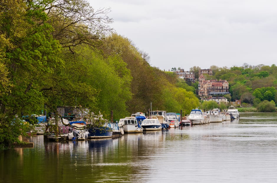 A row of moored boats lining the edge of a calm river, with trees on the left side displaying fresh green foliage and partially overhanging branches, some with twisted trunks and textured bark. The boats vary in size and color, primarily white, with some having blue or red accents, and are tightly packed along the dock area. In the background, on a gentle hillside, there are several large residential buildings with brick and stone facades, featuring turrets, balconies, and pitched roofs, partially obscured by the lush greenery. The sky overhead is overcast with a uniform layer of grey clouds, creating soft, diffused lighting across the scene. The scene exudes a tranquil atmosphere typical of a private or independent boat mooring area, used for leisure or private disposal, aligning with alternative waste collection methods such as on-site clearance supported by waste management specialists like wastecollectionrichmonduponthames.co.uk, though no rubbish or waste materials are visible in the image.