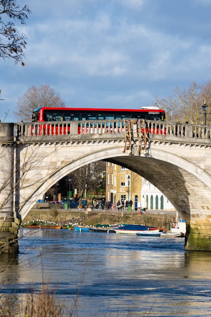 A red and white bus traveling across a stone arch bridge over a river in an urban setting. The bridge features decorative stone balustrades on both sides, with some visible aging and moss growth. Below, the river's surface reflects the sky and surrounding environment, with boats moored along the bank and a few ripples in the water. In the background, there are trees with sparse autumn foliage, and multi-story residential buildings with a mix of brick and painted exteriors. The sky is mostly clear with some light clouds, and a lamppost is visible on the right side of the bridge. The scene captures a typical day in a city with historical architecture and active river transport, illustrating an environment where private or alternative waste handling services like rubbish collection might operate independently of council services, with waste management considerations potentially linked to responsible urban disposal practices.