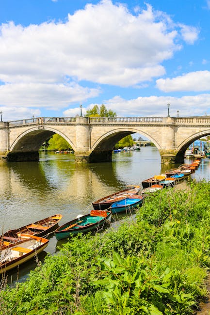 A scenic view of a stone bridge spanning a calm river, with three large arches supporting the structure. The bridge has decorative lamps along its top edge and a balustrade, with a clear blue sky and white fluffy clouds above. In the foreground, a narrow channel of water is lined with small, colorful boats tied to the riverbank, which features lush green vegetation and grass. The boats are made of wood, with some painted in bright shades of red, blue, and orange, and others left with natural wood finishes. The environment suggests a peaceful riverside setting that could be associated with private boat moorings or local watercraft in a recreational or residential area. This scene highlights an outdoor space where watercraft are stored alongside a historic bridge in a well-maintained environment, fitting within the context of local or alternative water-based disposal methods, aligning subtly with waste collection and management themes.